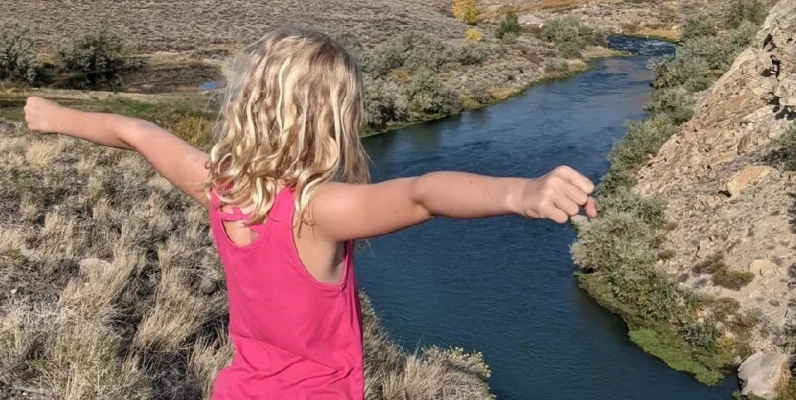 Nathan's daughter standing by cliff overlooking rive with arm out wide