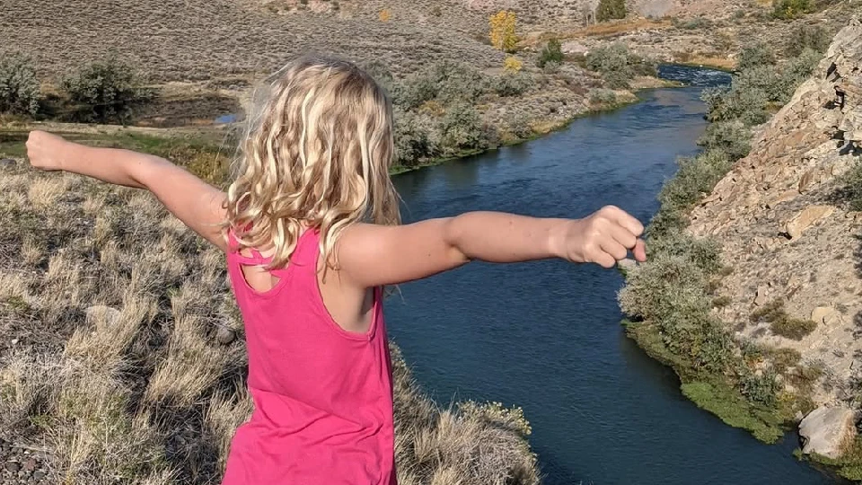 Nathan's daughter standing by cliff overlooking rive with arm out wide